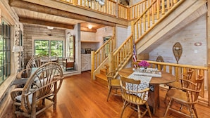 A rustic cabin interior with a wooden dining table, chairs, a staircase, and a balcony with a view of the outdoors. - Waterfront Cabin! Dock, Crab, Paddle, Kayak, Outdoor Movie, Karaoke, and More! (Heathsville)