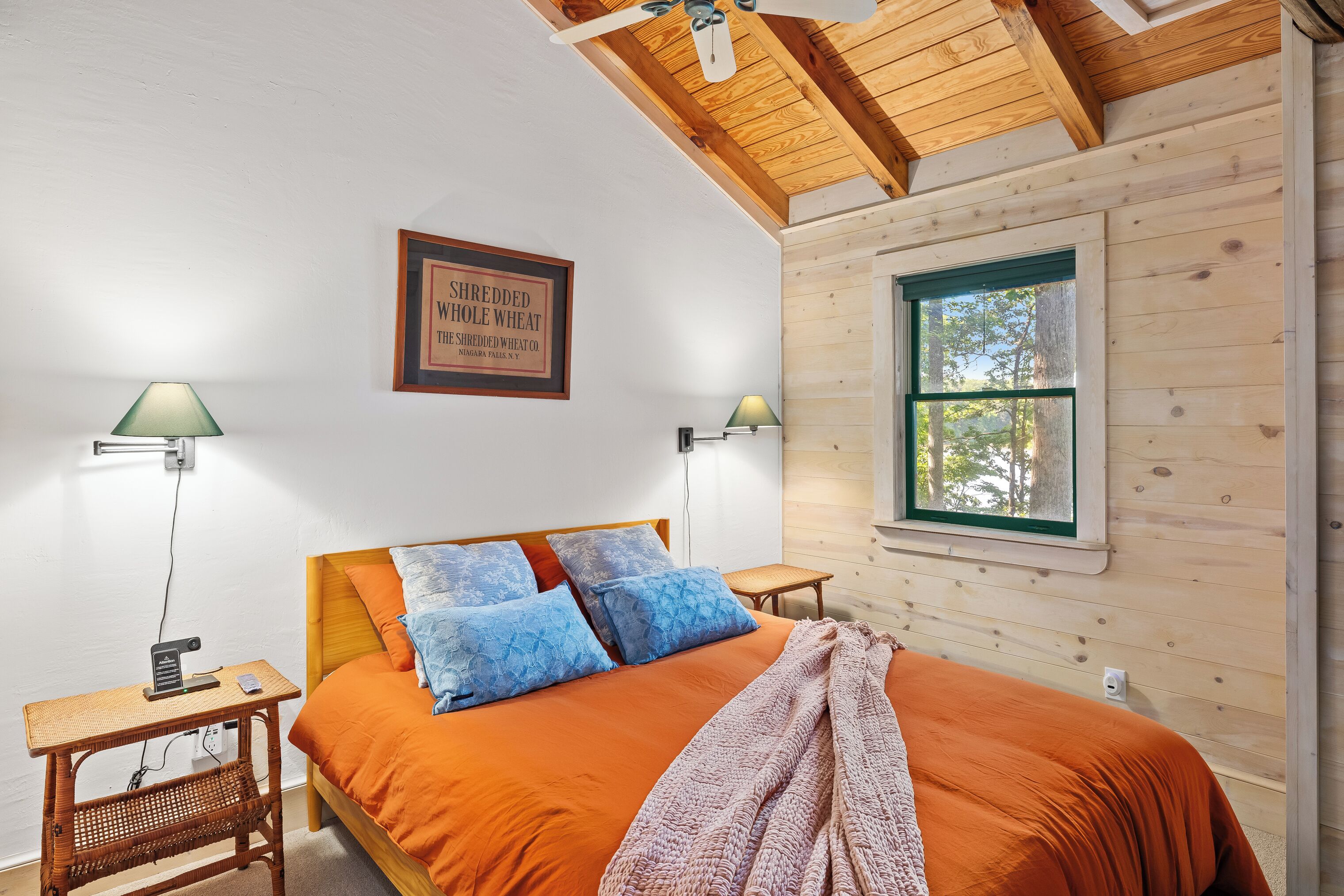 A bedroom with a wooden ceiling, a bed with orange bedding, two wall-mounted lamps, and a framed sign on the wall.
