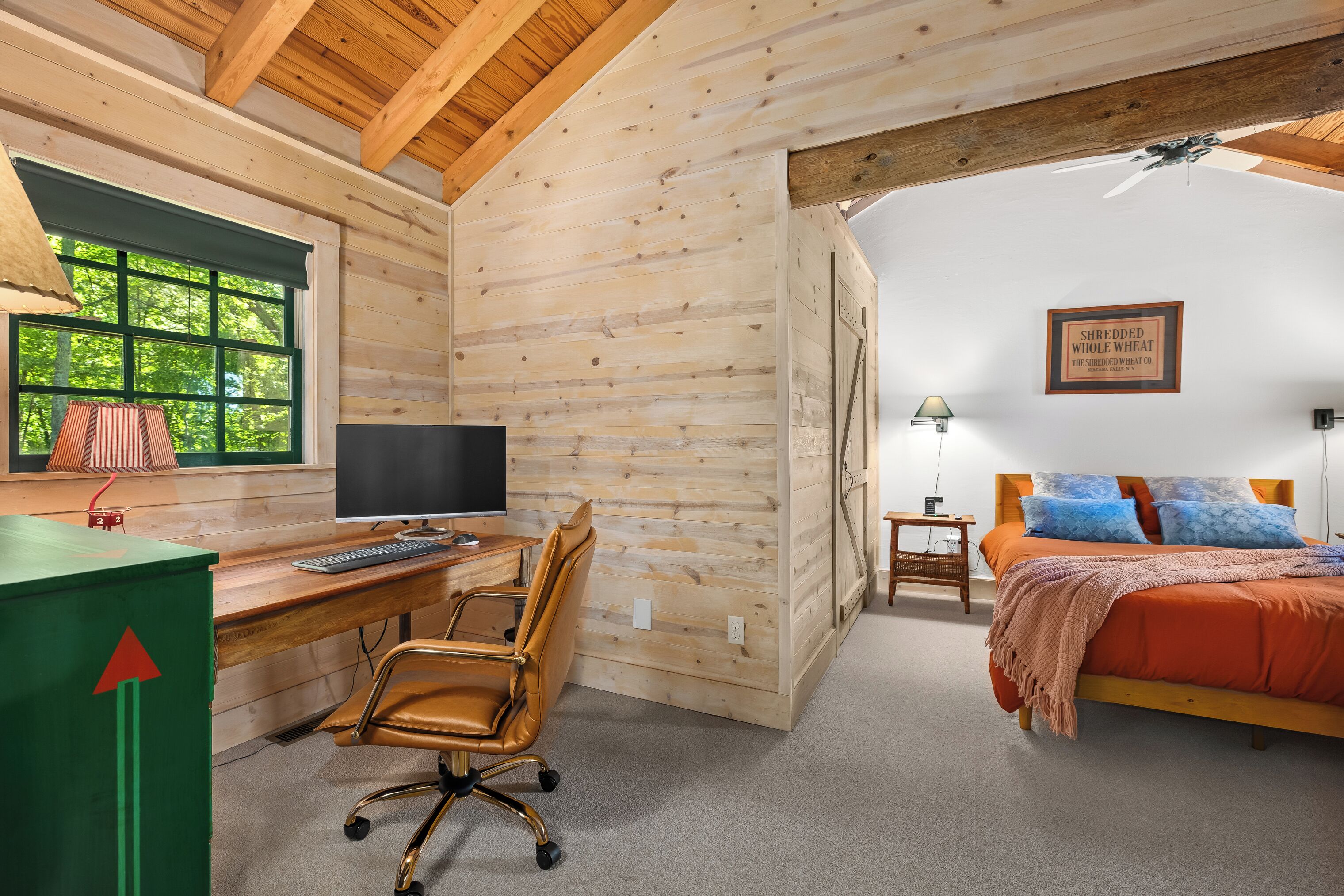 Interior de una cabaña con un escritorio de madera, computadora, silla y una cama con una manta roja.