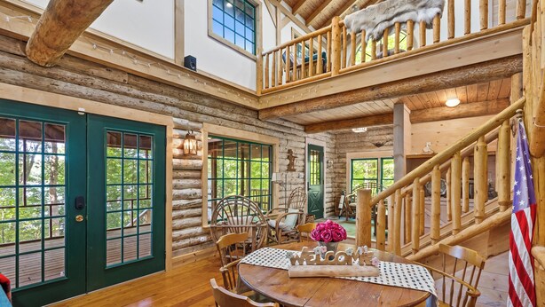A log cabin interior with a wooden dining table, chairs, a staircase, and a view of the outdoors. - Waterfront Cabin! Dock, Crab, Paddle, Kayak, Outdoor Movie, Karaoke, and More! (Heathsville)