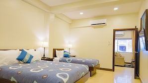 Habitación de hotel con dos camas, cada una con una colcha azul estampada y almohadas blancas. Cuenta con un ventilador de techo, un equipo de aire acondicionado y una puerta que da a un balcón.