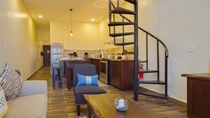 A spacious living area with a high ceiling, a wooden dining table, a sofa with patterned cushions, a kitchen in the background, and a spiral staircase leading to an upper level.