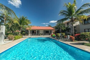 A rectangular swimming pool with clear blue water, surrounded by palm trees and a building with a red roof.