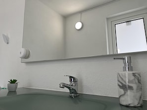 Une salle de bain avec un mur blanc, un miroir rond, un lavabo vert, un robinet argenté et un distributeur de savon en marbre.