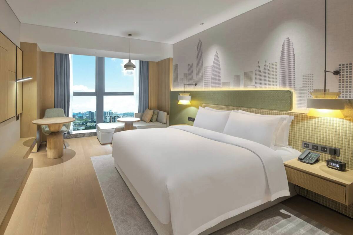A modern hotel room with a large bed, a television, a dining table, and a city view.