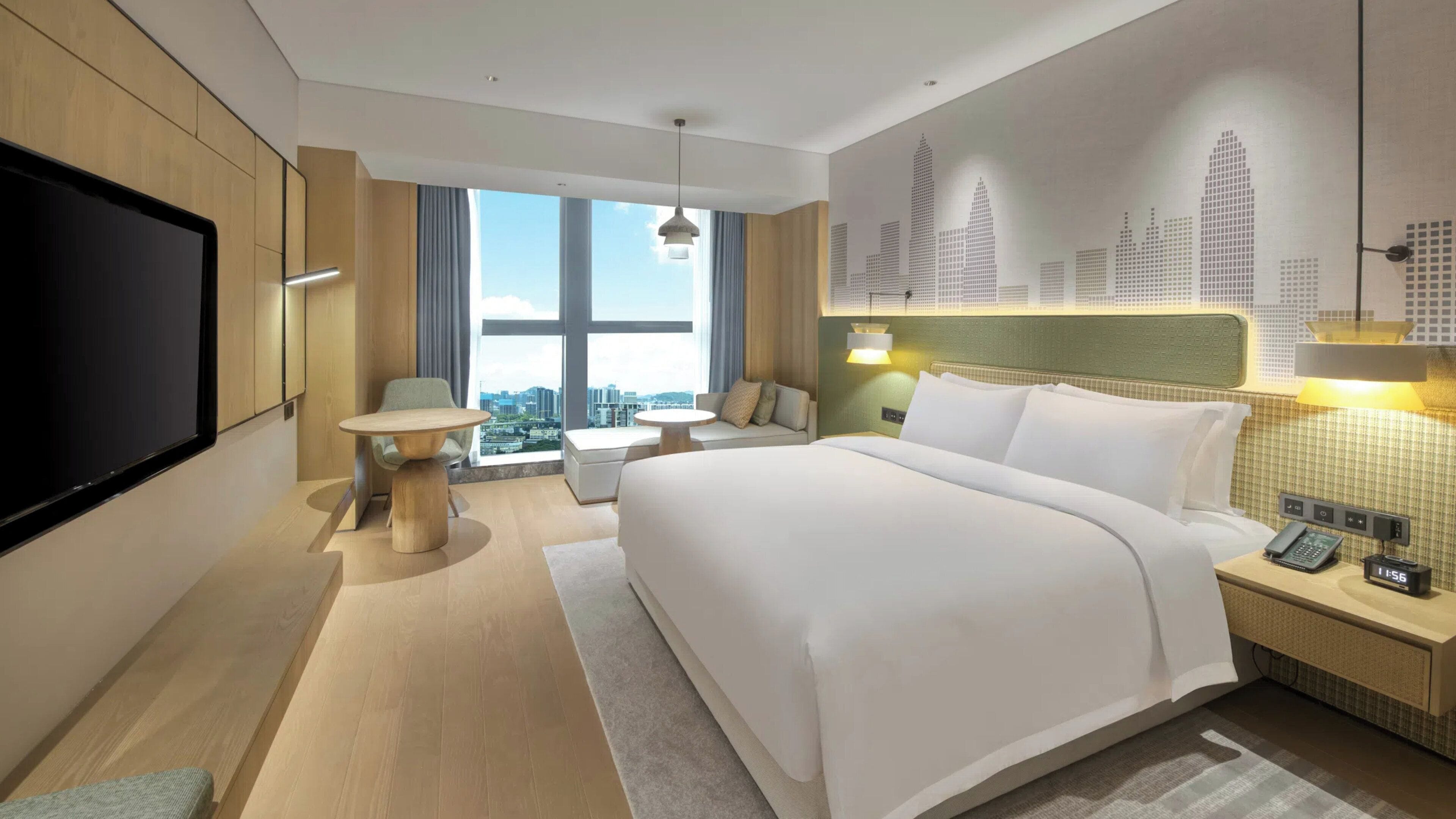 A modern hotel room with a large bed, a television, a dining table, and a city view.