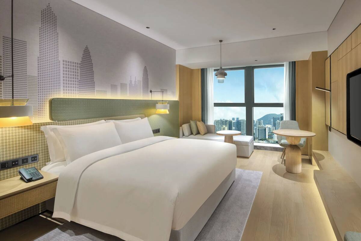 A modern hotel room with a large bed, a TV, a city view, and a small table.