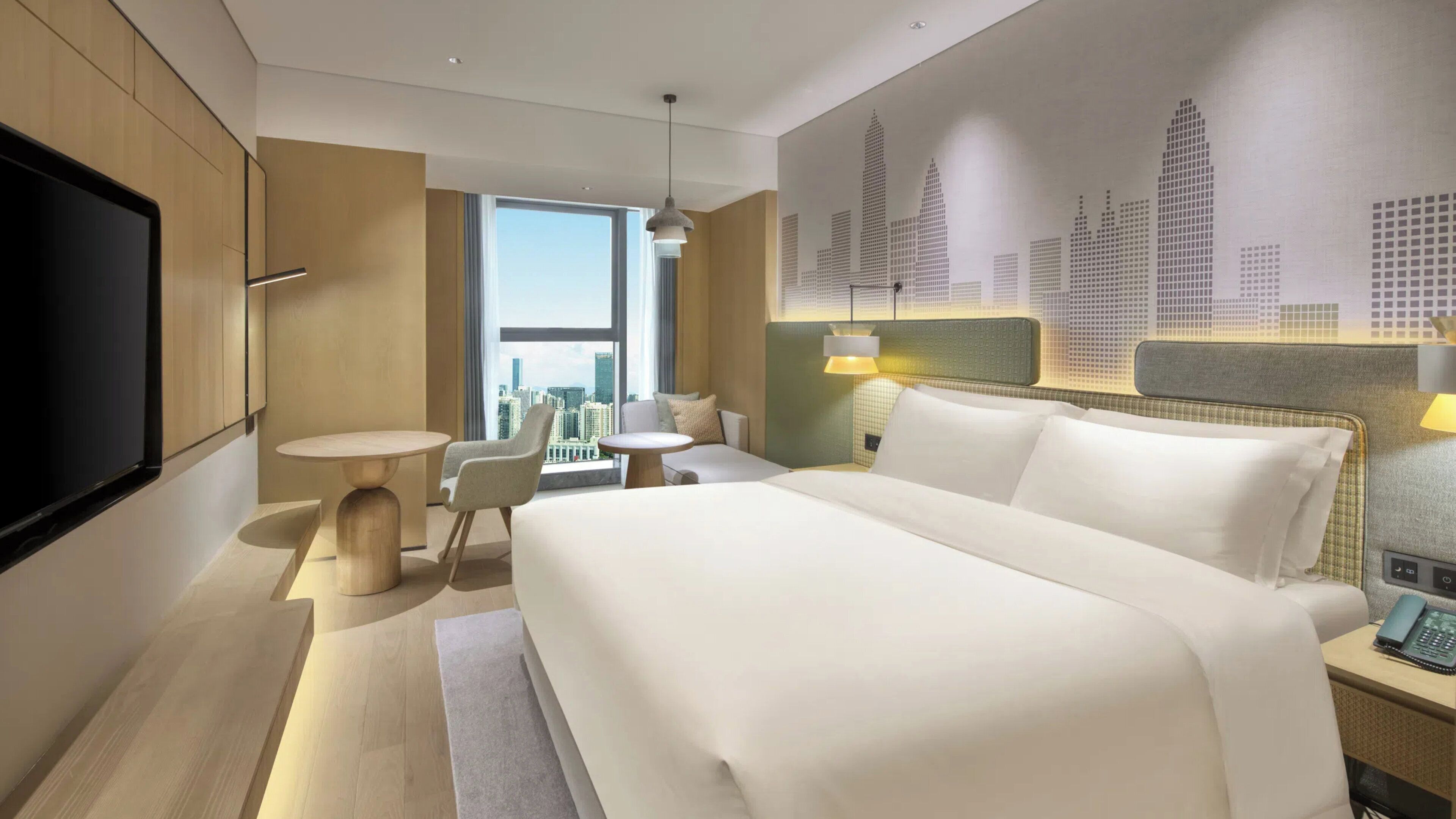 Ein modernes Hotelzimmer mit einem großen Bett, einem Fernseher, einem Schreibtisch und Blick auf die Stadt.