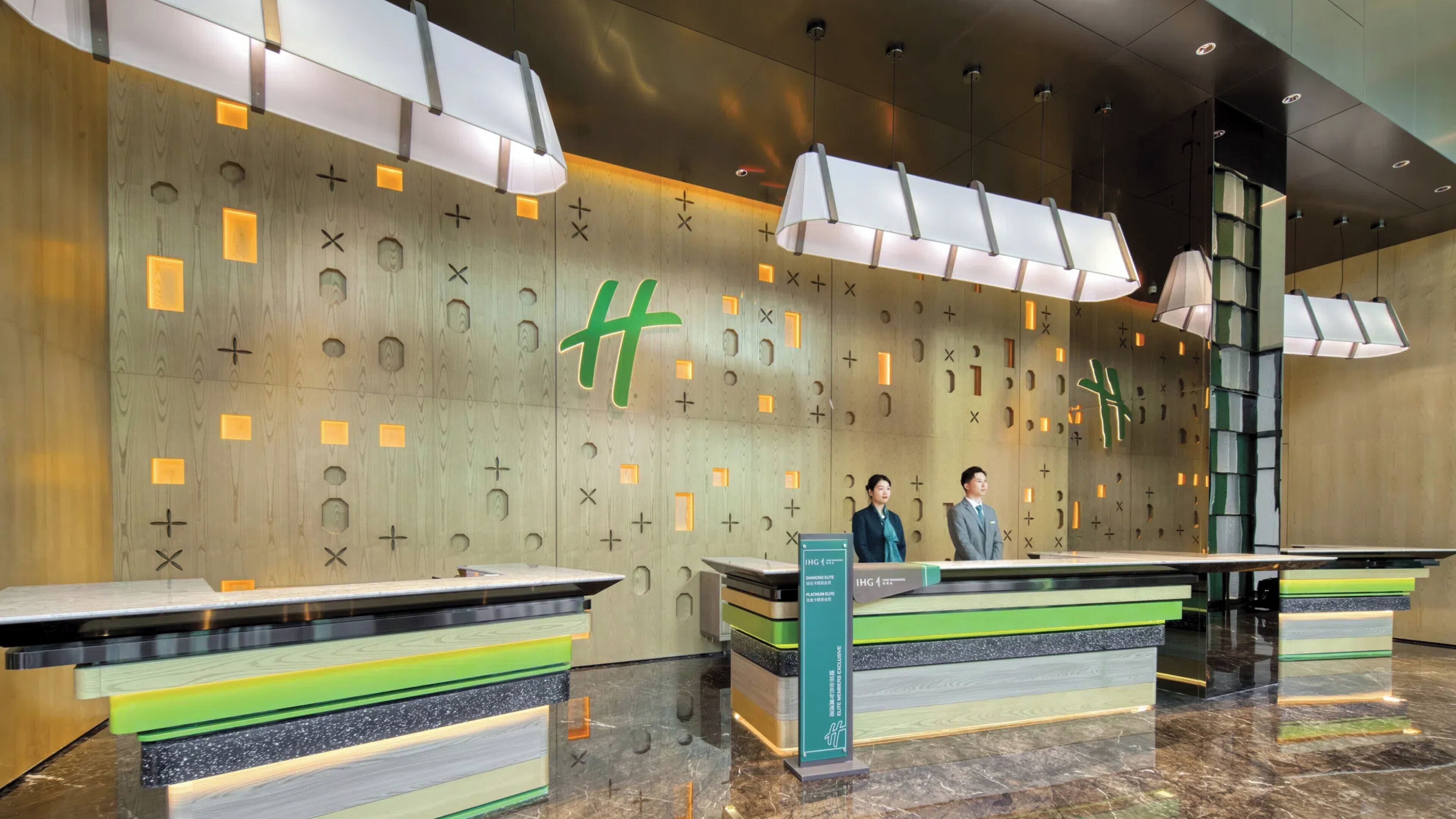 A hotel reception area with a modern design, featuring a large wall with geometric patterns and a prominent green 'H' logo. There are reception desks with glass tops and a sign indicating 'IHG Diamond Lounge'.
