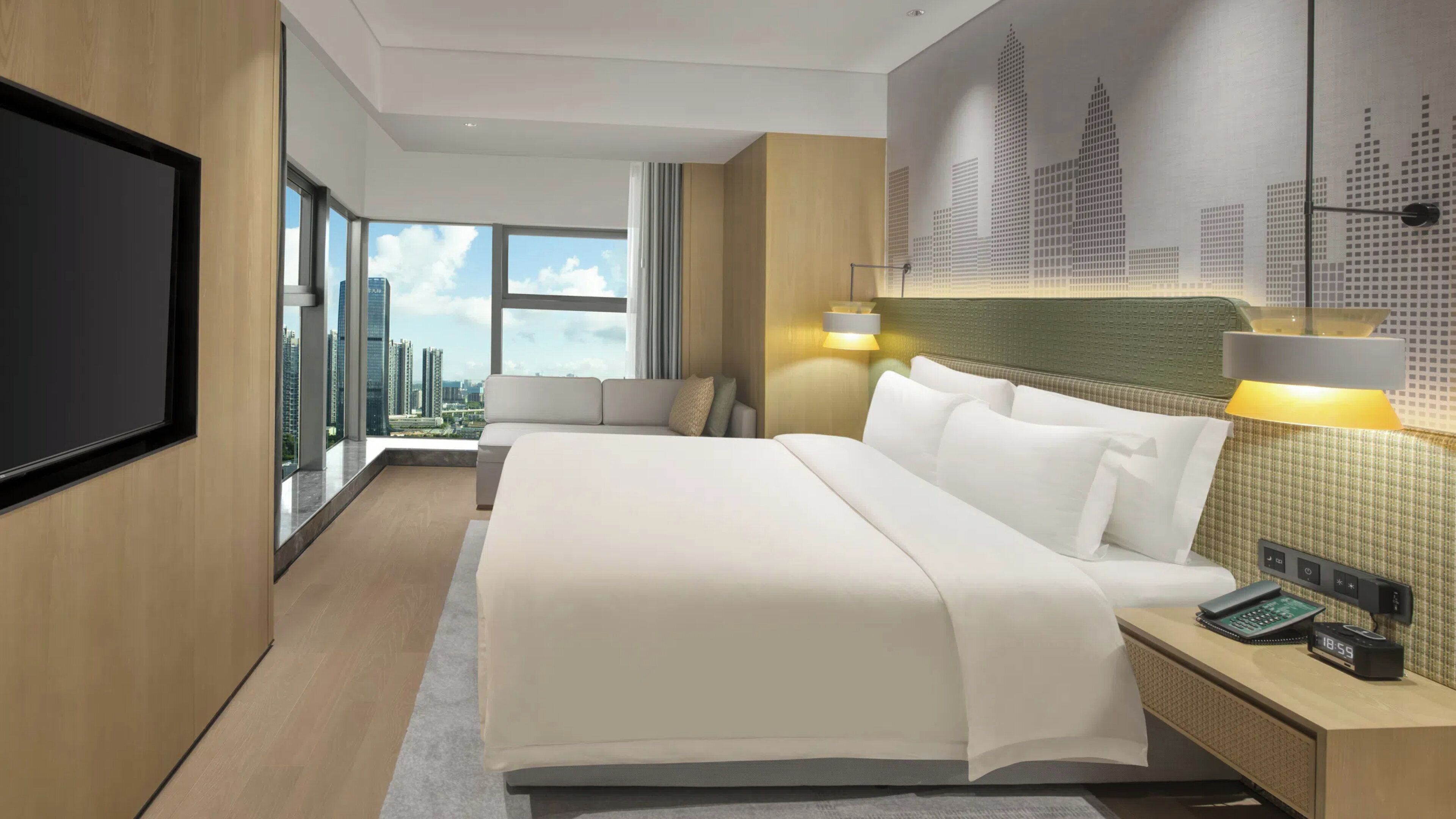 Een moderne hotelkamer met een groot bed, uitzicht op de stad, een flatscreen-tv en een bureau met telefoon en wekker.