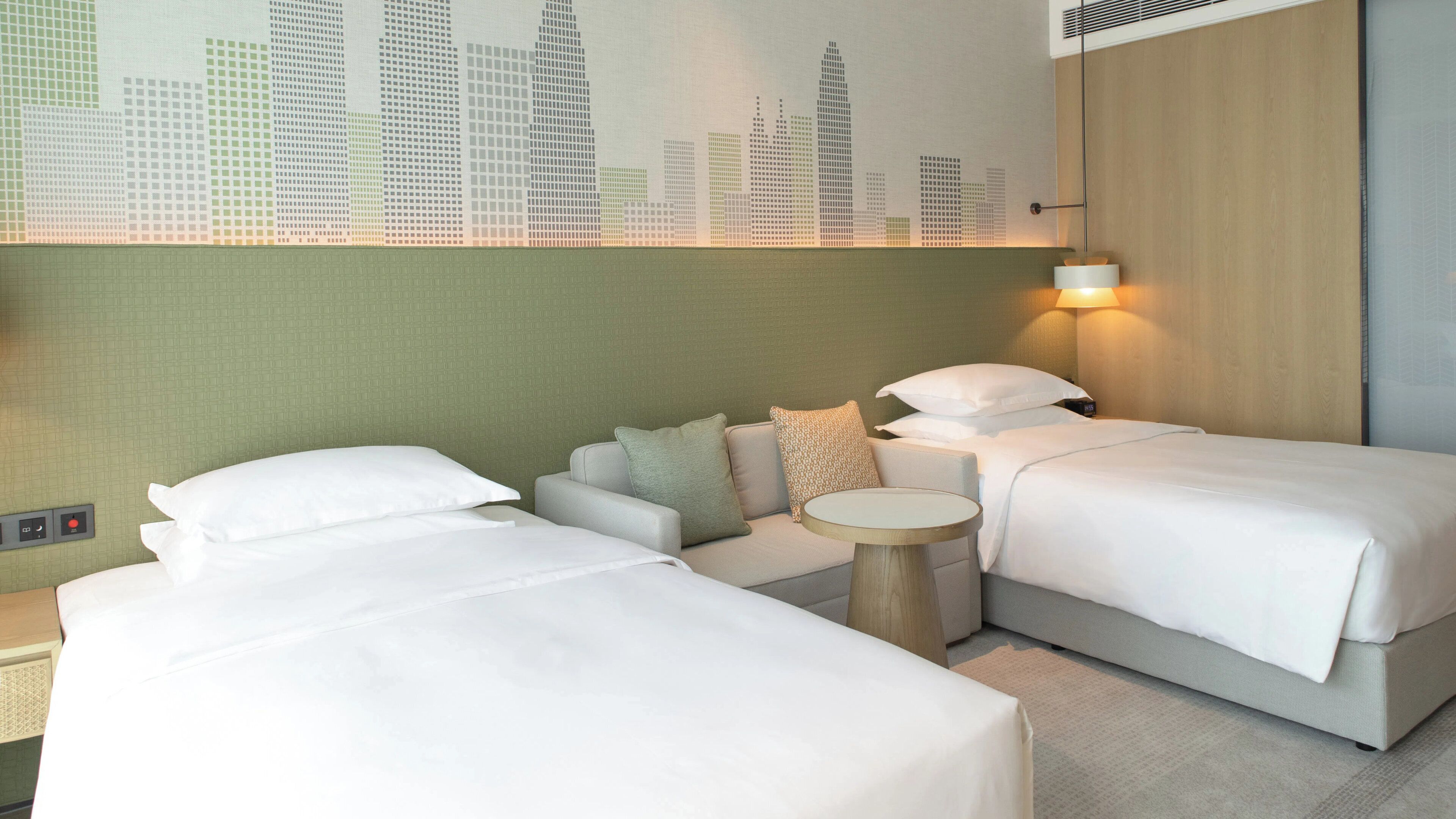 A hotel room with two beds, a sofa, a round table, and a cityscape mural on the wall.