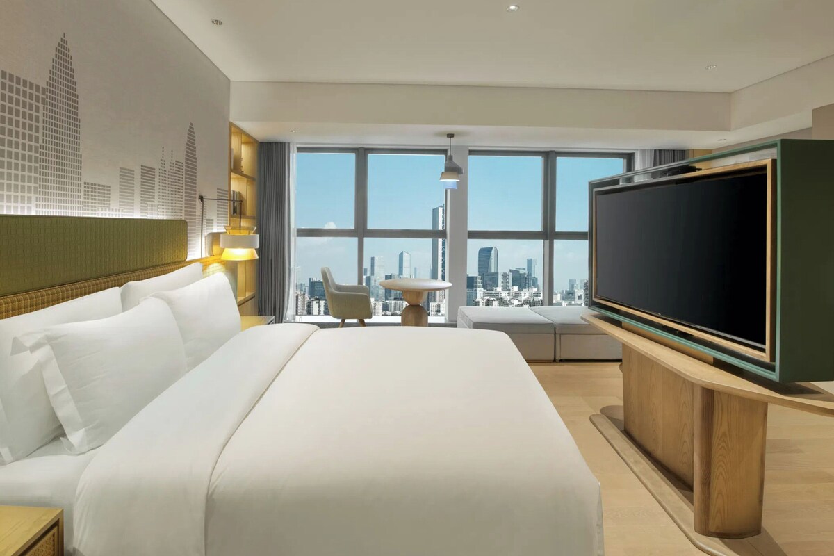 Een hotelkamer met een groot bed, een televisie, een bureau met een lamp, een stoel en uitzicht op een stadsbeeld door grote ramen.