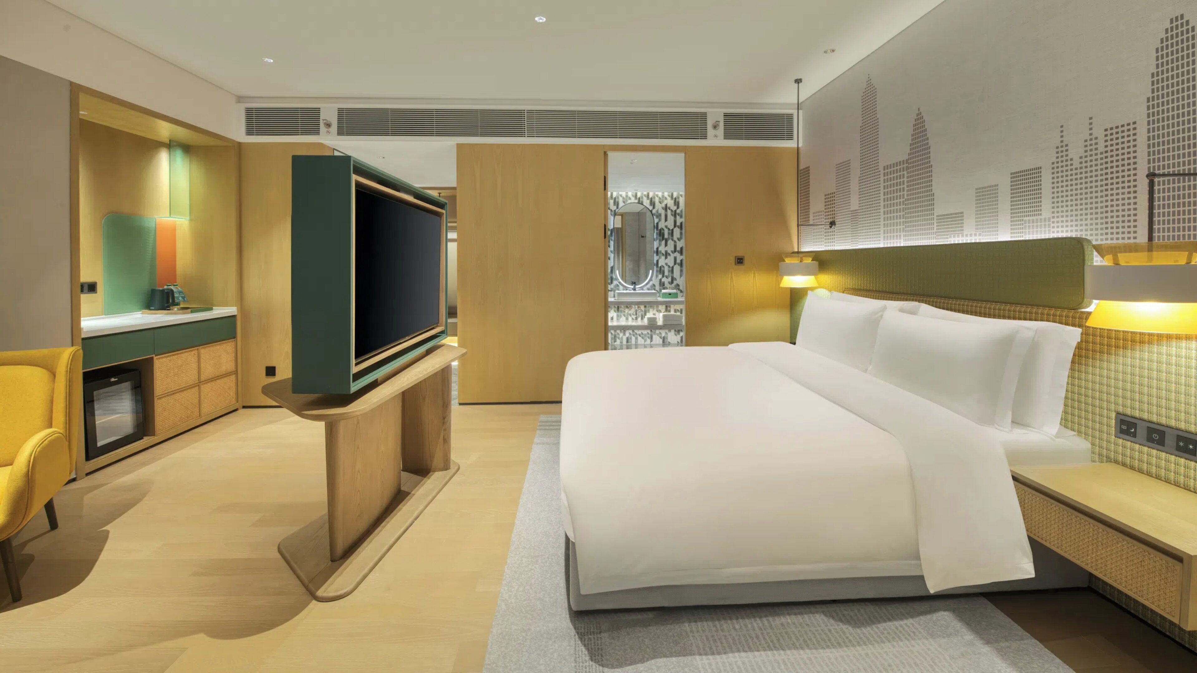 Una habitación de hotel moderna con una cama grande, un televisor sobre un soporte de madera, un escritorio con una lámpara y un mural de un paisaje urbano en la pared.