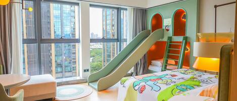 A hotel room with a large window, a bed with a colorful bedspread, a desk, a chair, a small table, and a children's play area with a slide and ladder.