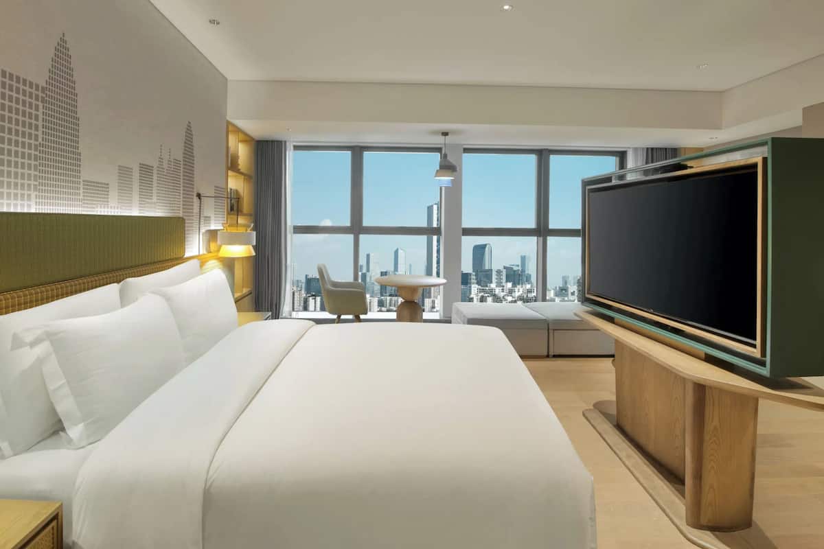 A hotel room with a large bed, a television, a desk with a lamp, a chair, and a view of a cityscape through large windows.
