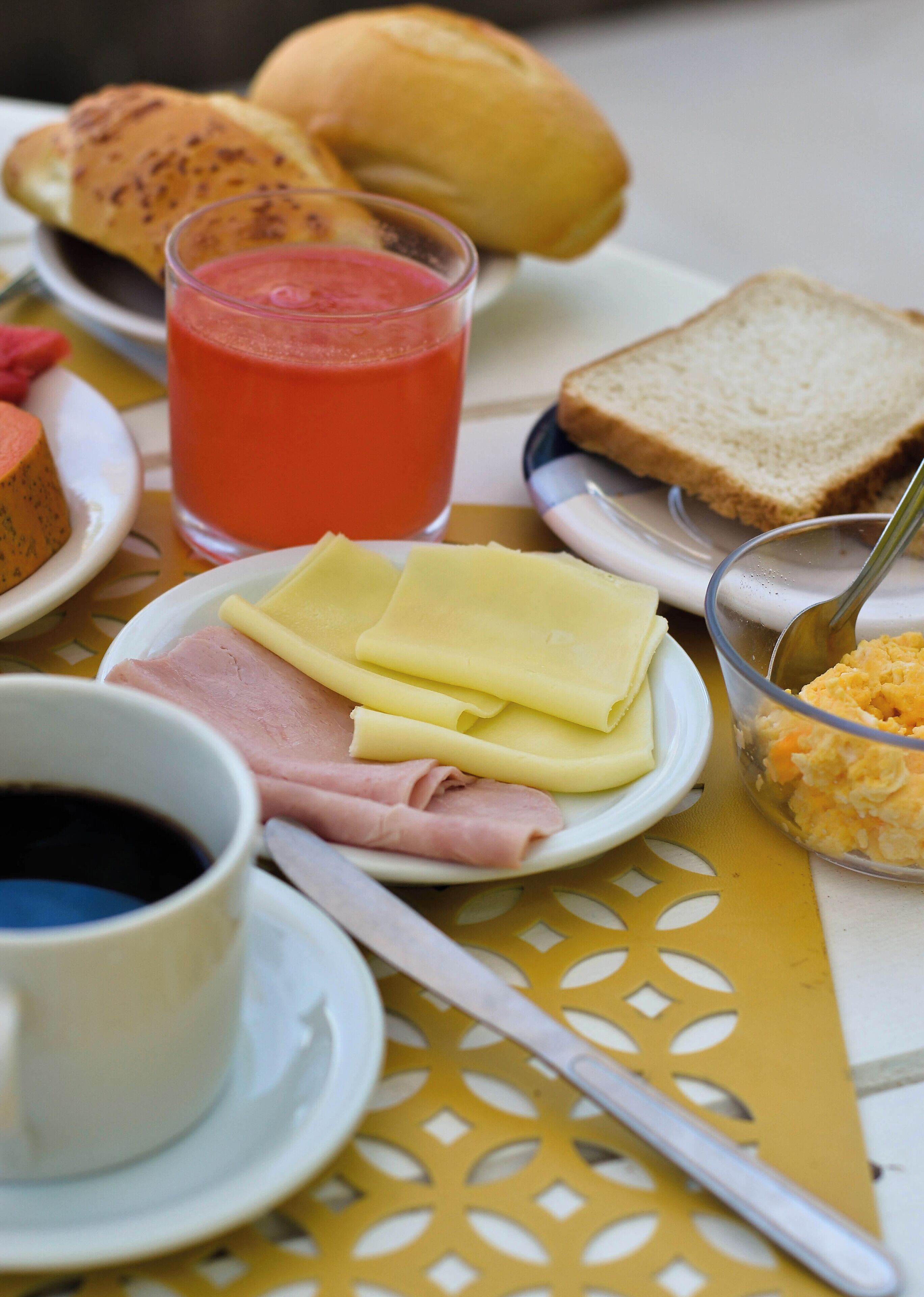 A breakfast setting with bread, cheese, ham, a glass of juice, and a cup of coffee on a table with a yellow tablecloth.