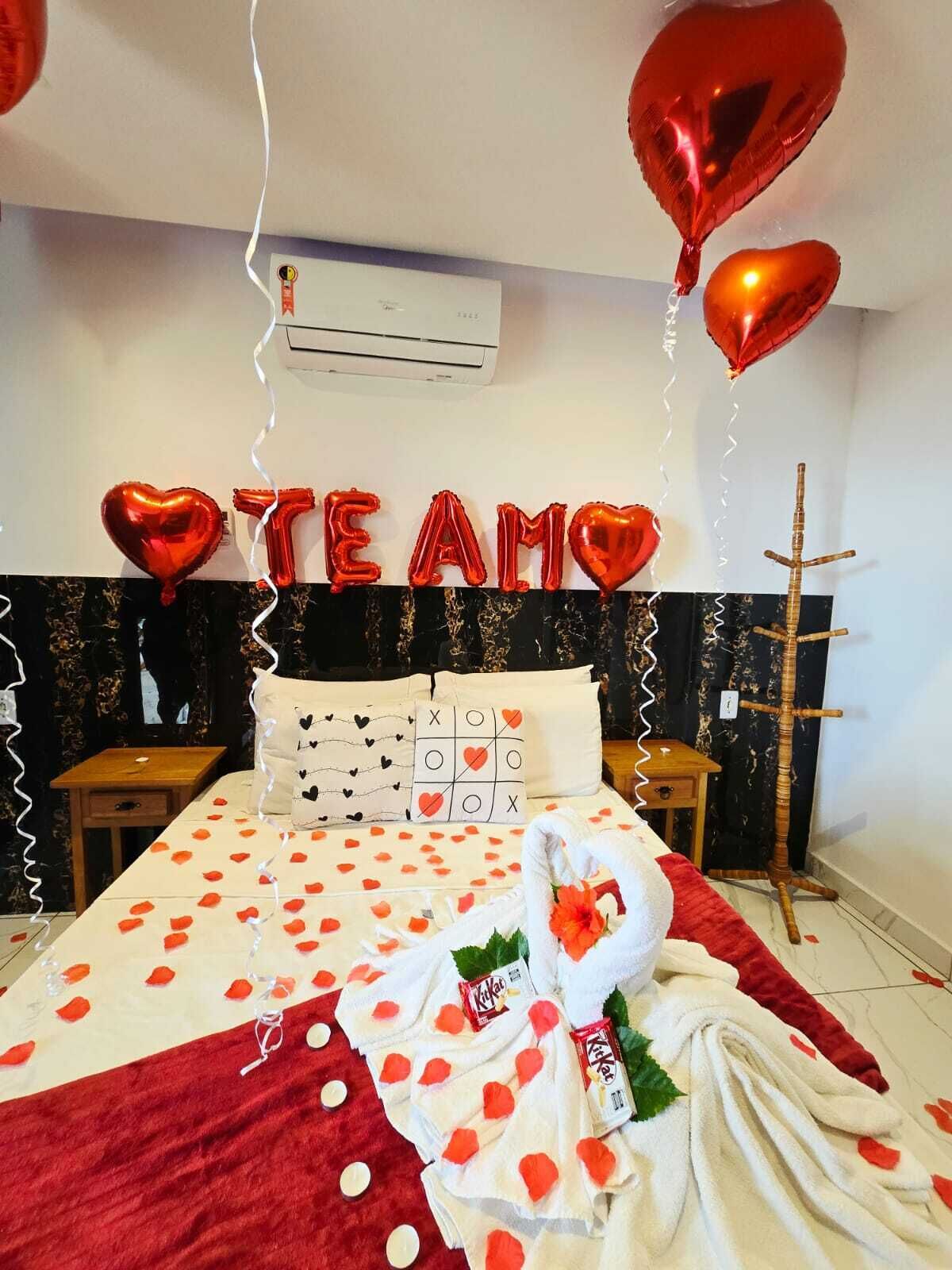 A hotel room with a bed, pillows, a bedside table, a wall-mounted air conditioner, and heart-shaped balloons spelling out 'TEAM'.