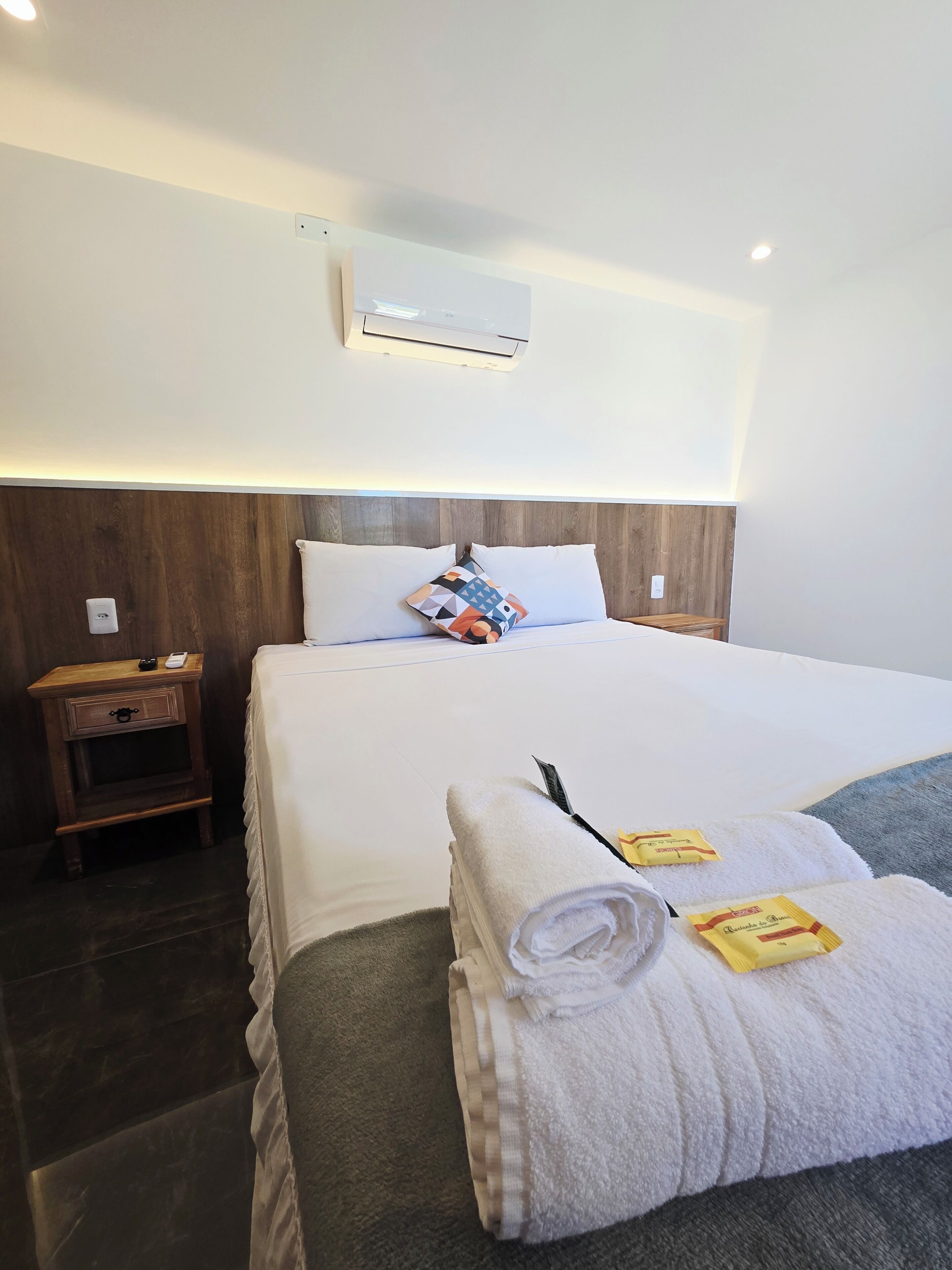 A hotel room with a neatly made bed, a wooden nightstand, a wall-mounted air conditioner, and a bedside table with a lamp.