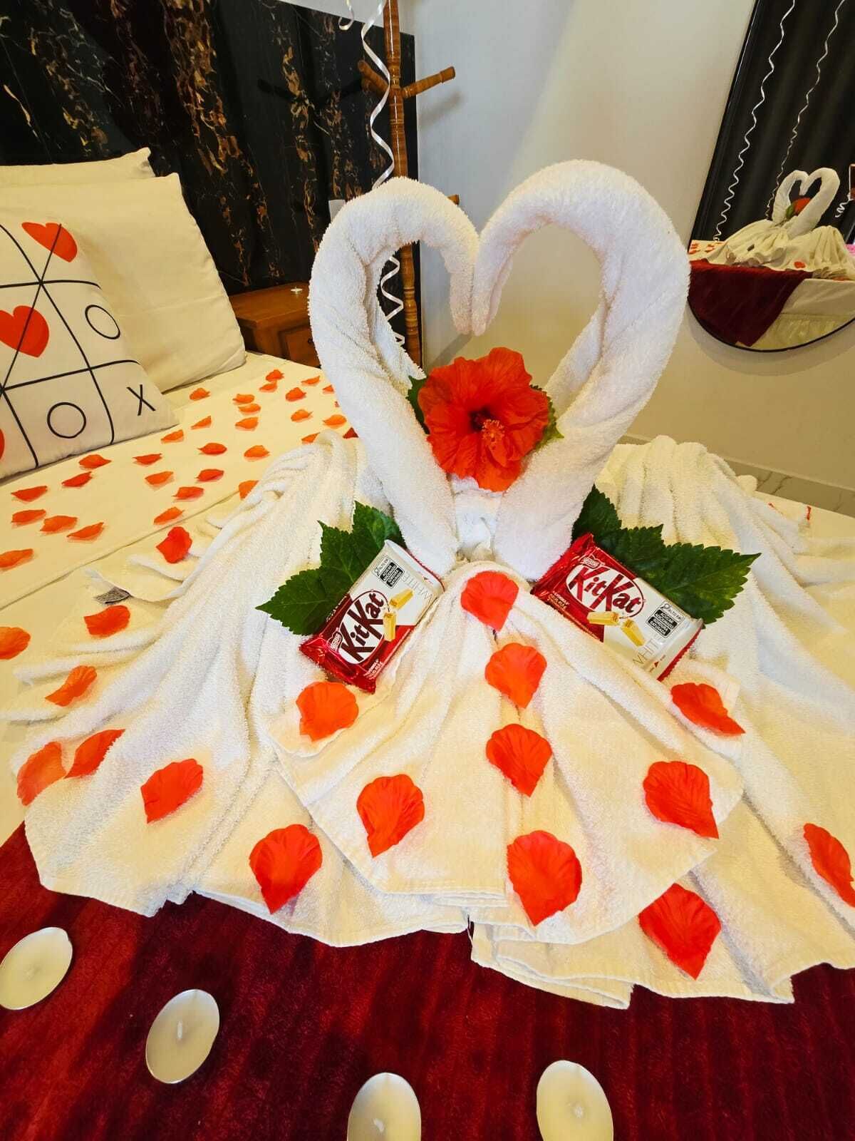 A room with a bed covered in a white towel with red flower petals, a KitKat chocolate bar, and a red flower centerpiece.