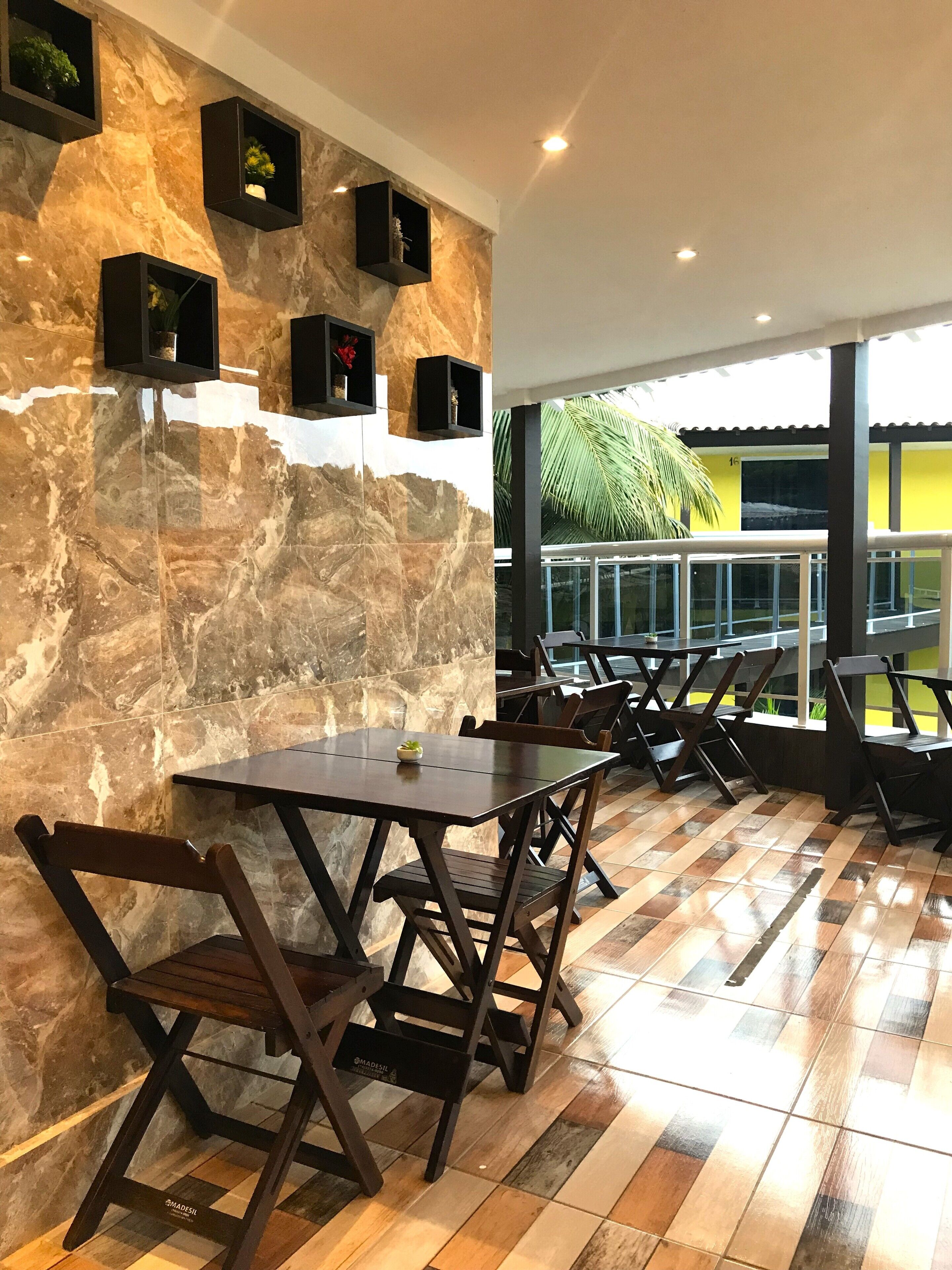 A restaurant with marble walls, black shelves with plants, wooden tables and chairs, and a view of the outdoors.
