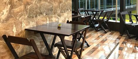 A restaurant with marble walls, black shelves with plants, wooden tables and chairs, and a view of the outdoors.