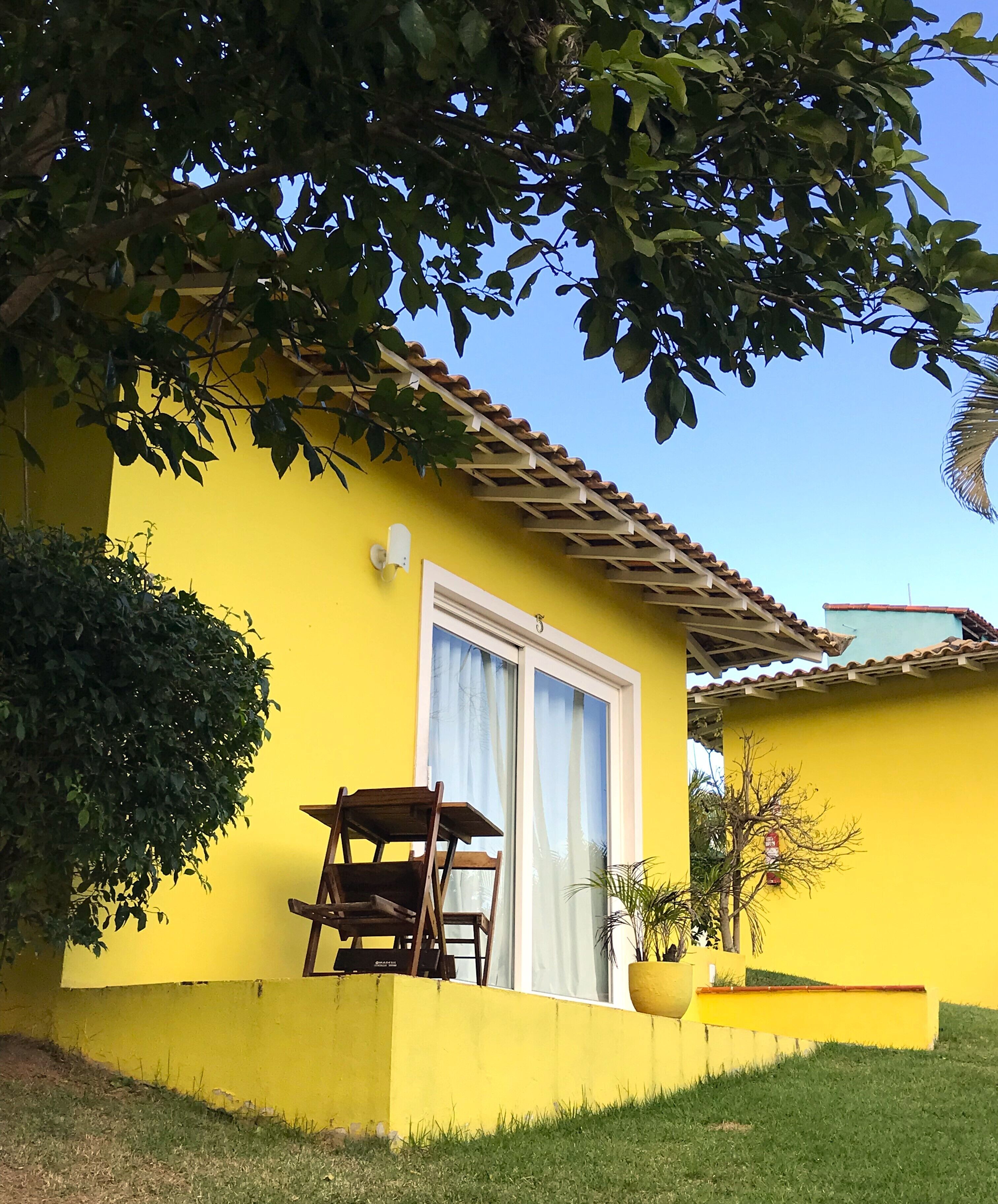 A yellow house with a tiled roof, a window with a white curtain, and a wooden chair outside.