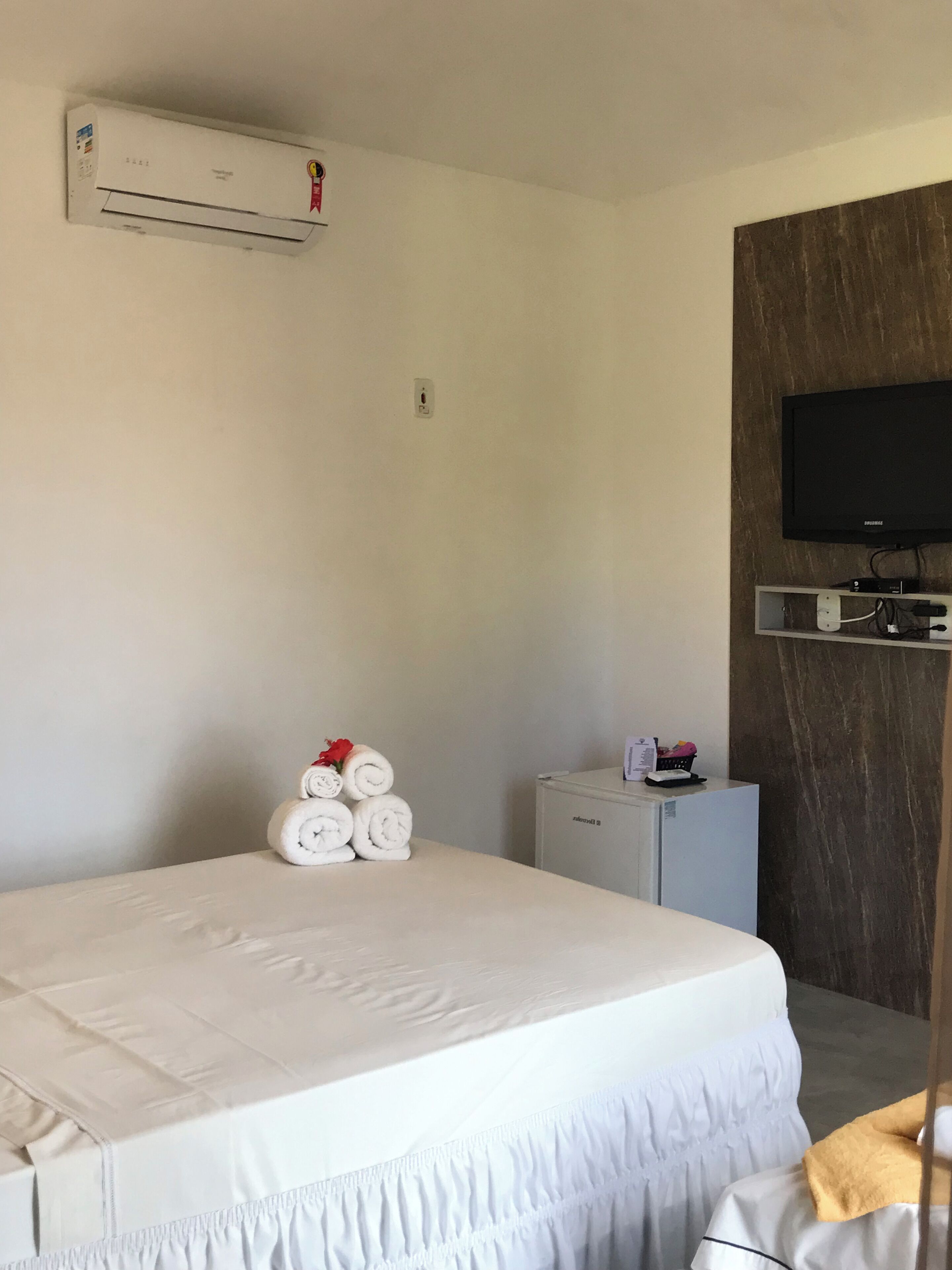 A hotel room with a bed, an air conditioner, a television, a small refrigerator, and a wall-mounted TV.