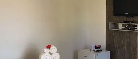 A hotel room with a bed, an air conditioner, a television, a small refrigerator, and a wall-mounted TV.