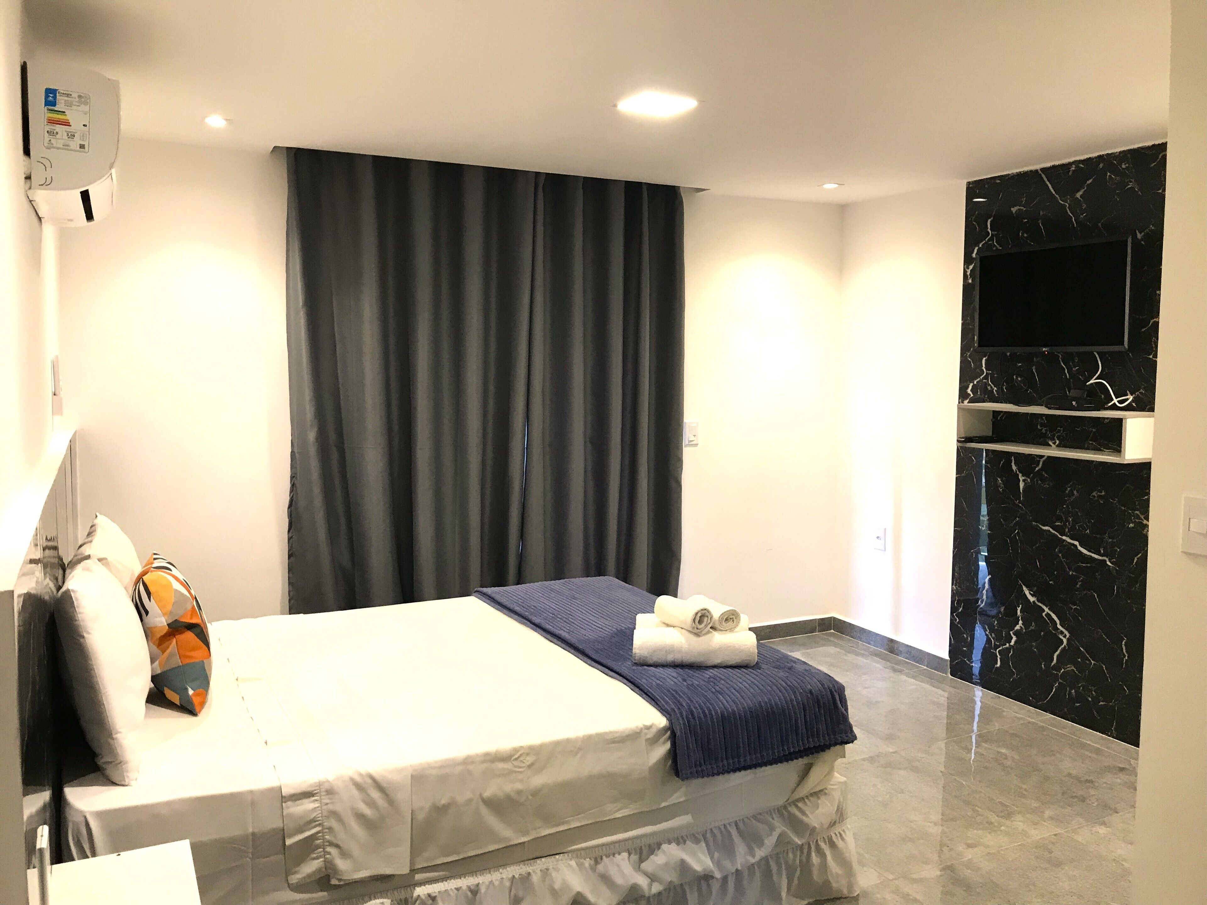 A hotel room with a bed, a flat-screen TV mounted on a black marble wall, and a window with dark curtains.