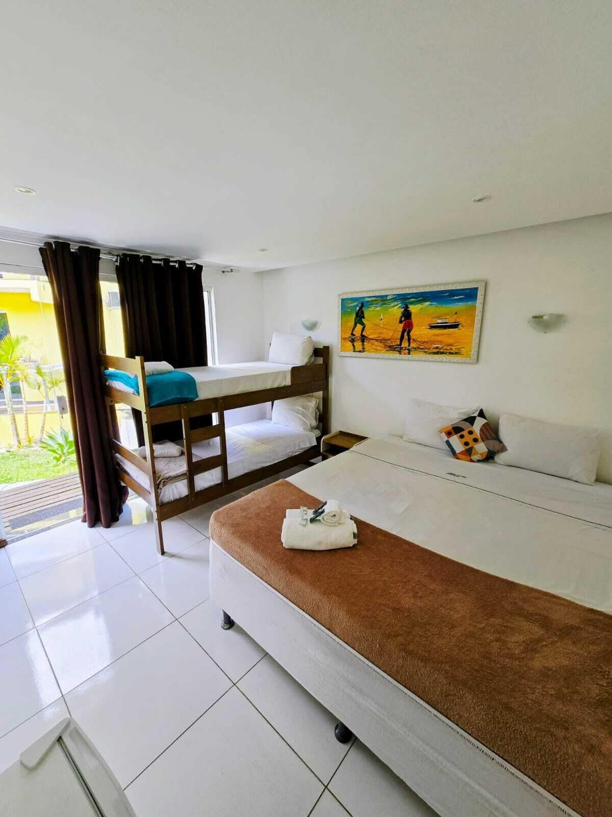 A hotel room with a bunk bed, a single bed, a painting of a beach scene, and a view of the outdoors.