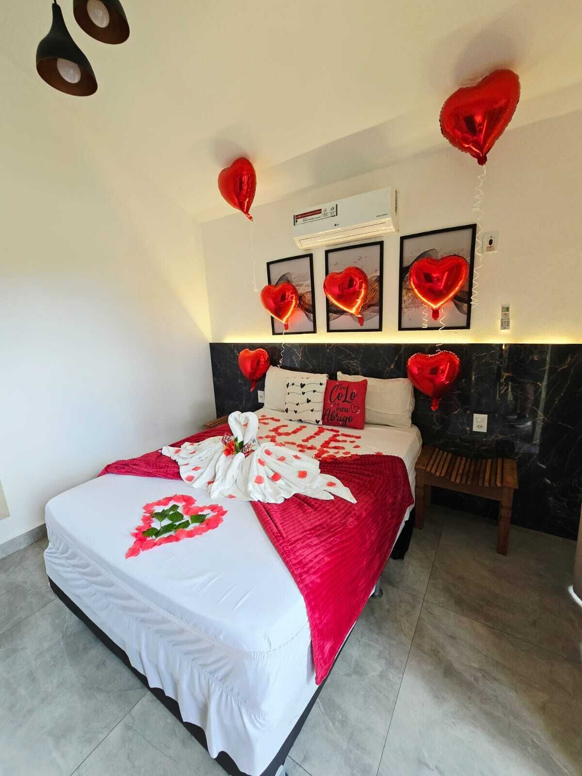 Una cama con una colcha roja y blanca, globos con forma de corazón y un ramo de flores.