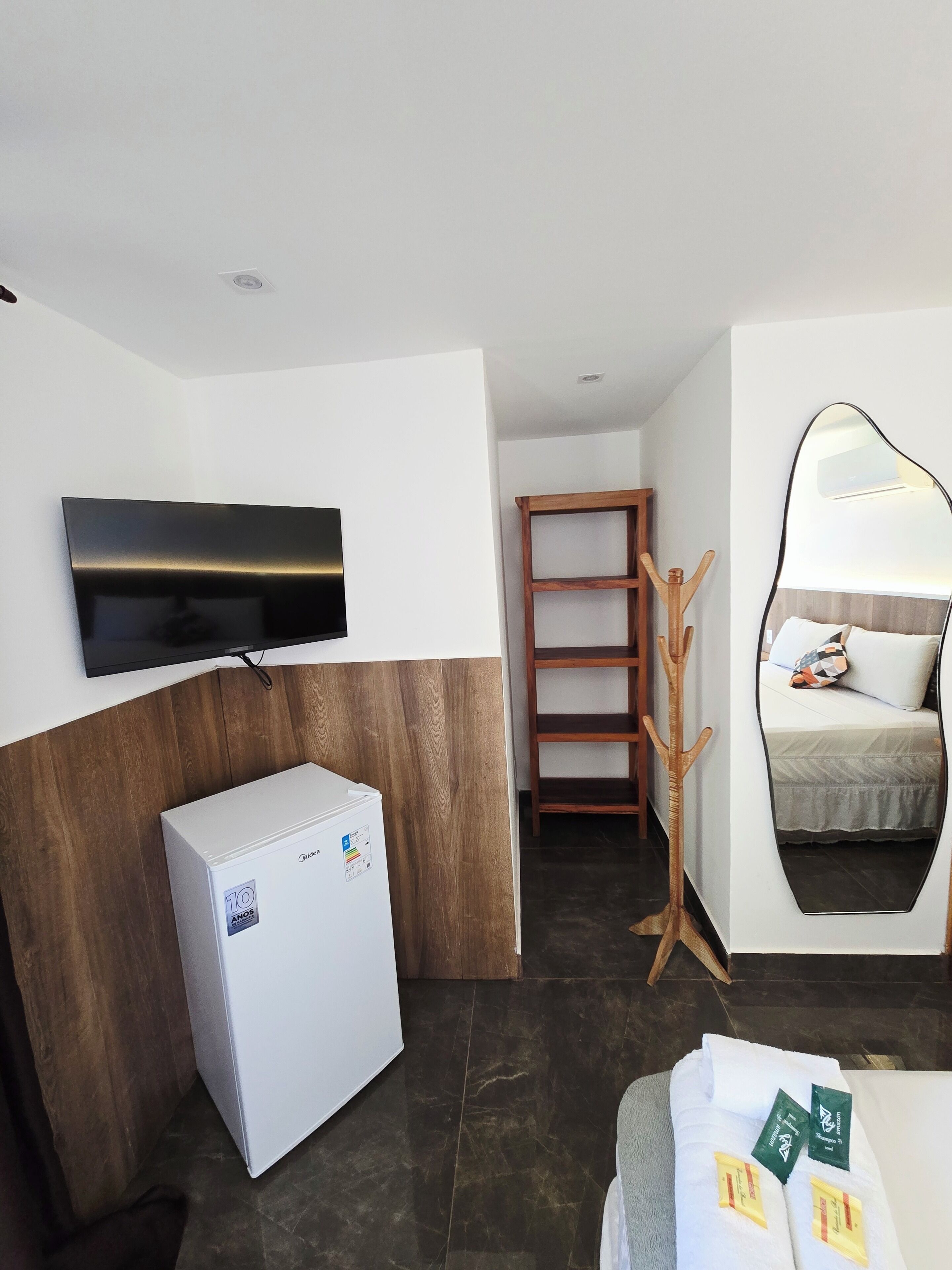 A modern hotel room with a flat-screen TV, a minibar, a wooden coat rack, a bed with a mirror, and a tiled floor.