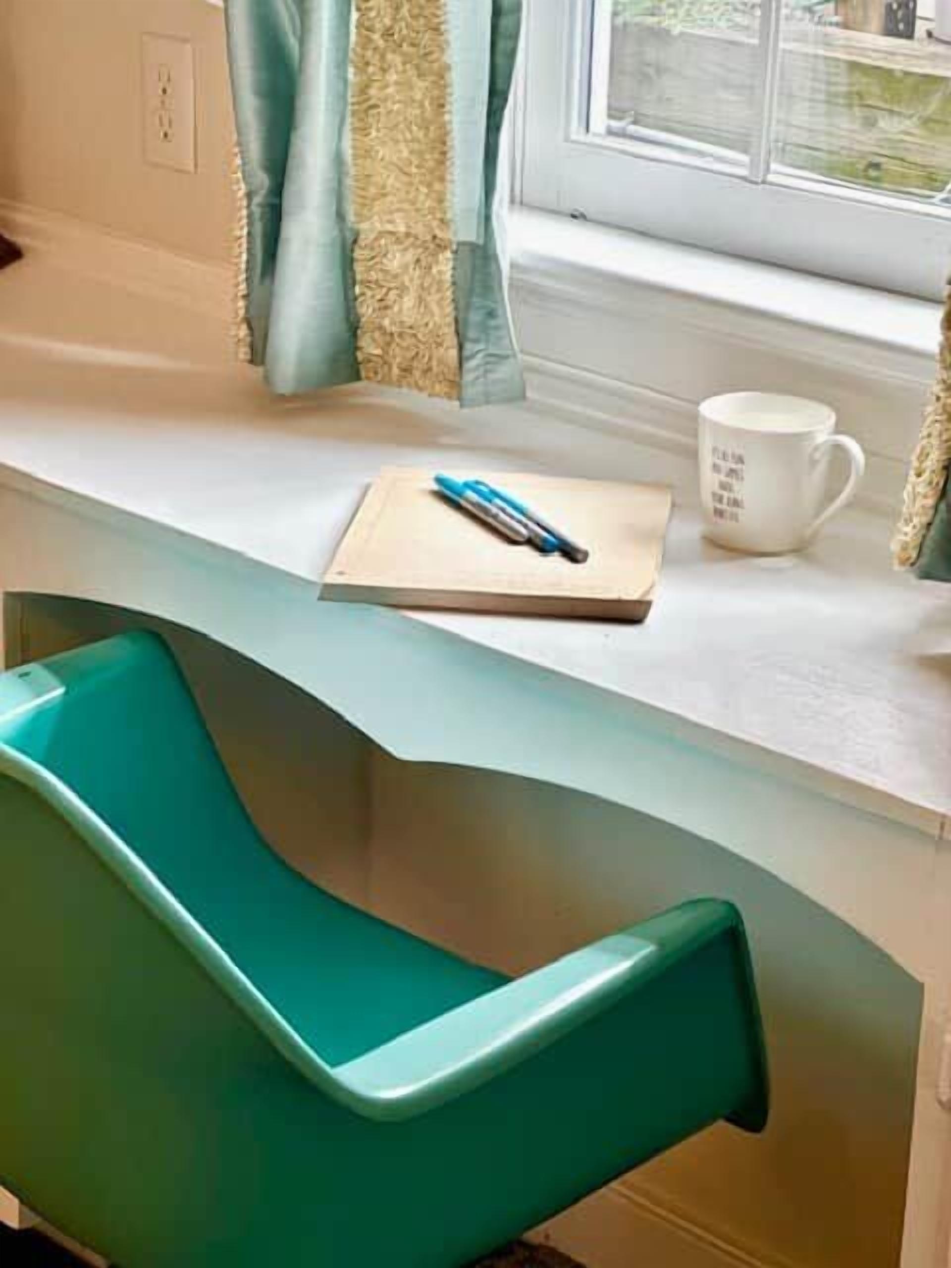 Una silla color turquesa junto a un escritorio blanco, con un cuaderno, un bolígrafo y una taza.