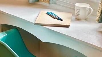 Una silla color turquesa junto a un escritorio blanco, con un cuaderno, un bolígrafo y una taza.