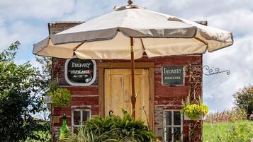 A rustic wooden building with a large umbrella, surrounded by greenery and a clear sky.