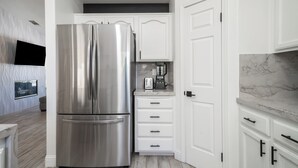 A modern kitchen with white cabinetry, a stainless steel refrigerator, a marble countertop, and a wall-mounted clock.