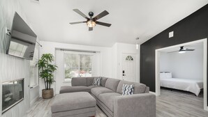 A modern living room with a sectional sofa, a ceiling fan, a large window, and a door leading to a bedroom.