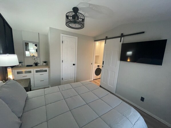 A bedroom with a bed, a dresser, a mirror, a door, a washing machine, and a TV mounted on the wall.