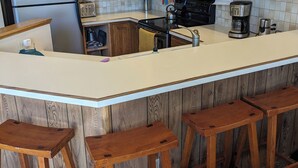 Une cuisine avec des meubles en bois, un réfrigérateur en acier inoxydable, un four à micro-ondes, une cuisinière et une machine à café. Il y a quatre tabourets en bois au comptoir.