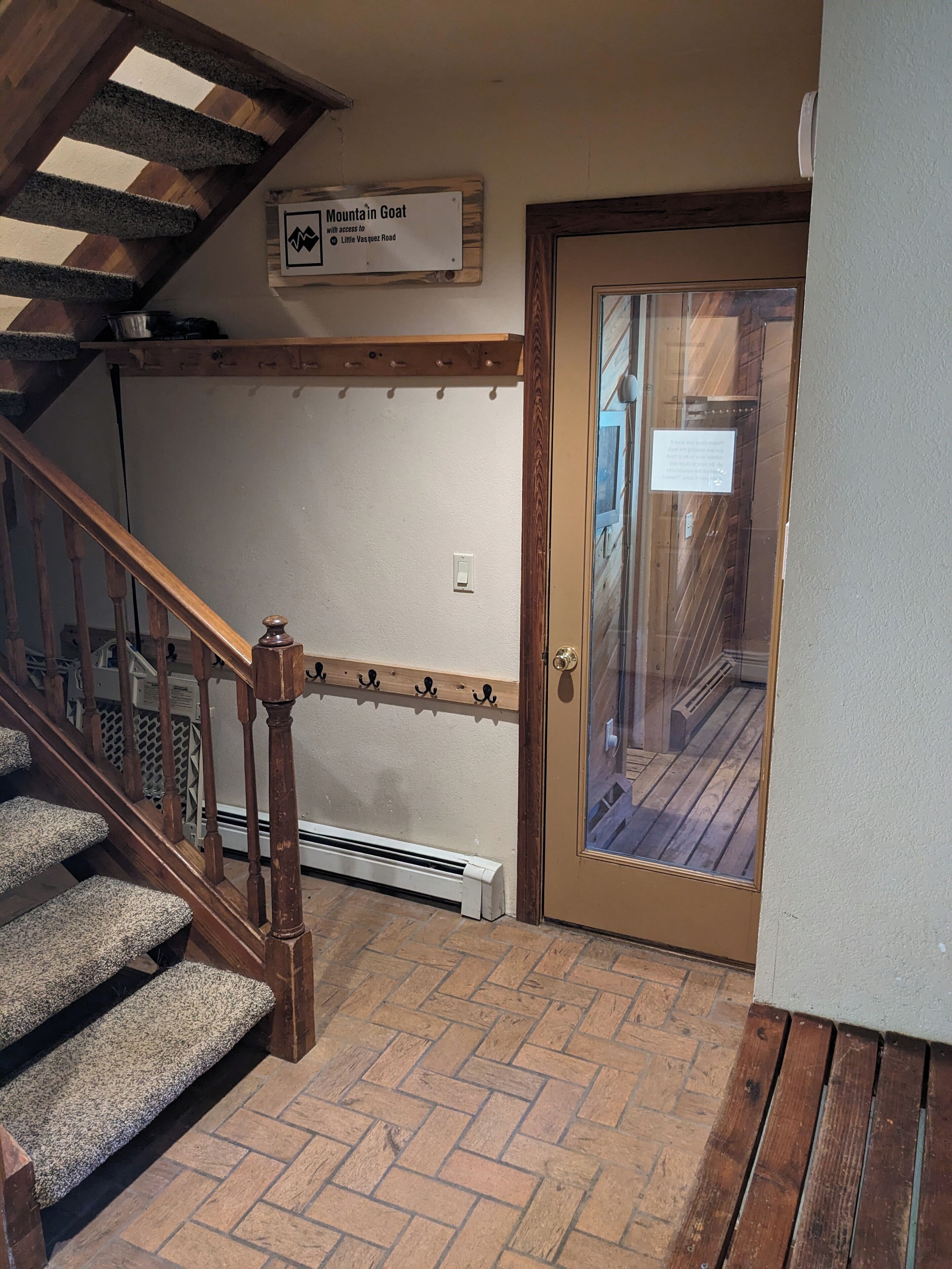 A staircase with wooden railings and carpeted steps leading to a door with a glass panel. A sign above the door reads "Mountain Goat with access to Little Vasquez Road."