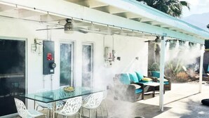An outdoor patio with a glass table and chairs, a covered seating area with cushions, and a visible fire extinguisher. - Palms Spring Doering Vista Estate (Palm Springs)