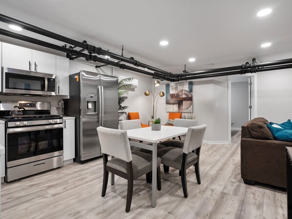 A modern kitchen with stainless steel appliances, a dining area with a white table and grey chairs, and a brown sofa in the living area.