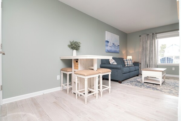 A living room with a light-colored wooden floor, a blue sofa, a small white table, and a decorative plant. - Walk to the Beach - Ocean Block - 1st Floor Condo (Dewey Beach)