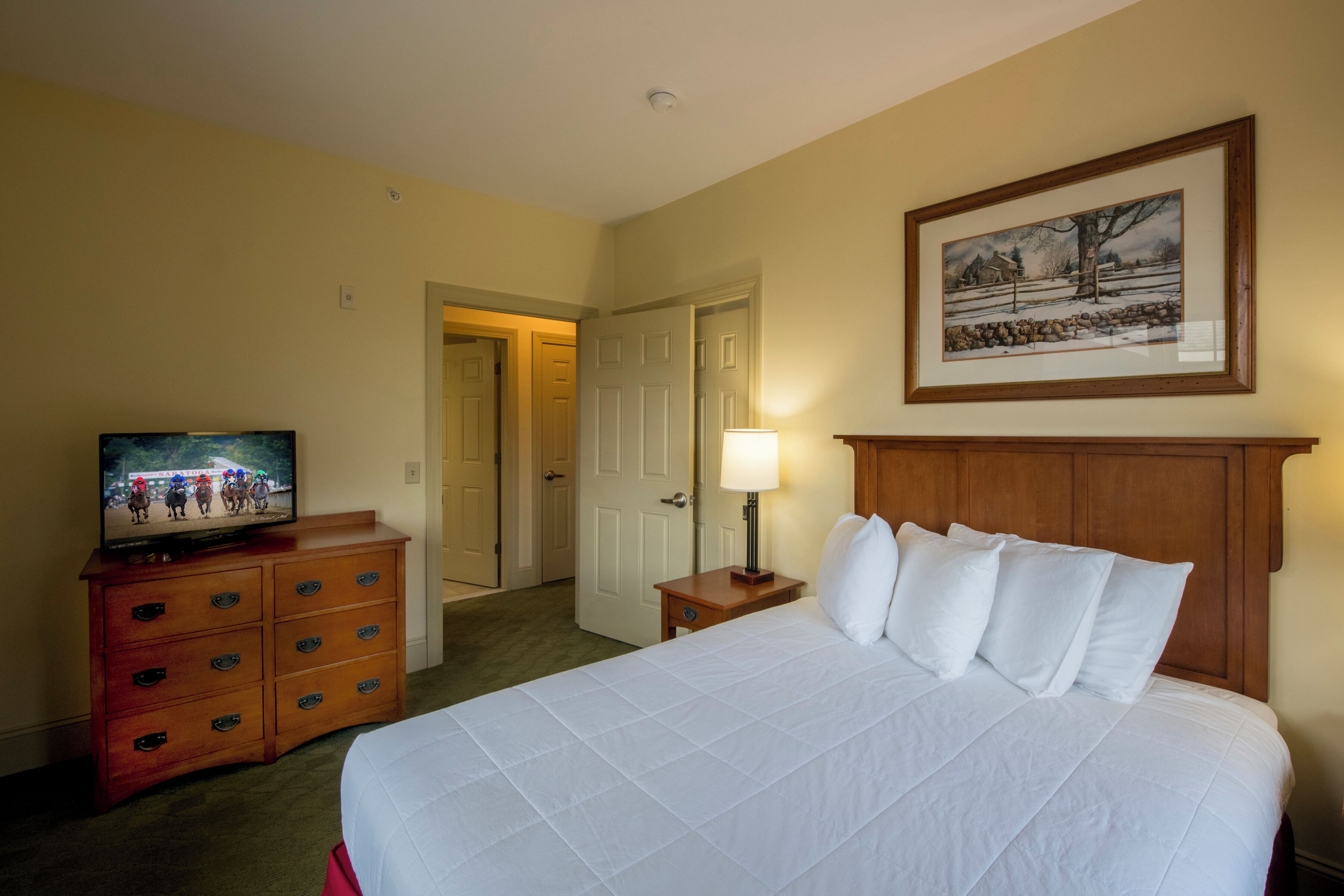 Una habitación de hotel con una cama grande, un aparador de madera, un televisor, un cuadro enmarcado en la pared y una puerta que da a otra habitación.