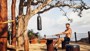 Un hombre se ejercita con un saco de boxeo al aire libre, rodeado de construcciones de madera y suelo arenoso.