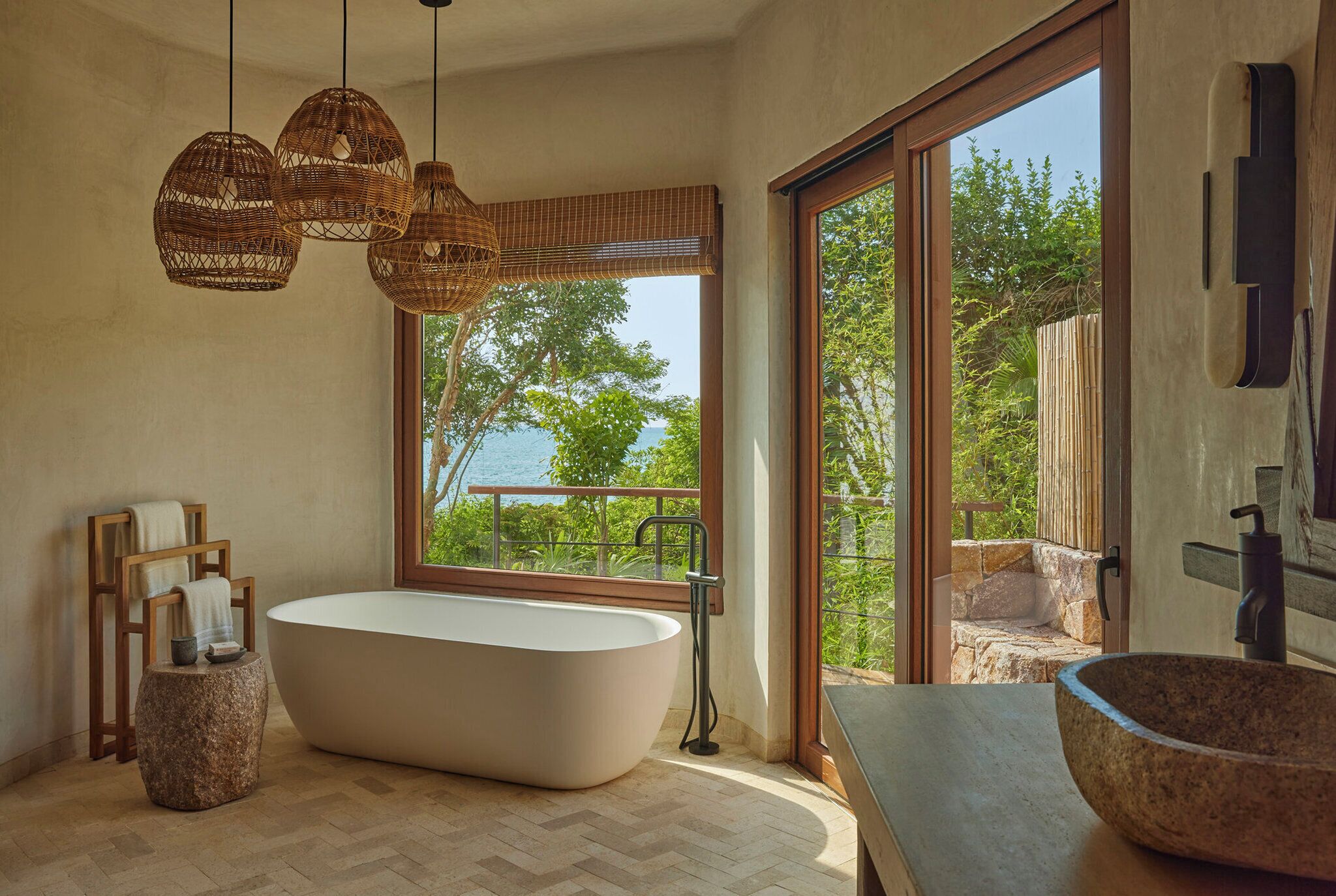 Un baño moderno con una bañera independiente, un amplio ventanal con vistas a la vegetación y un cuerpo de agua, y un lavabo de piedra.