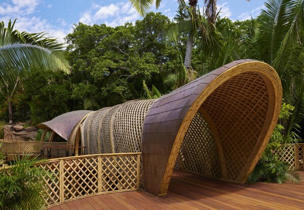 Una estructura grande, curva y sin techo, con tejido de techo, rodeada de exuberante follaje y una terraza de madera.