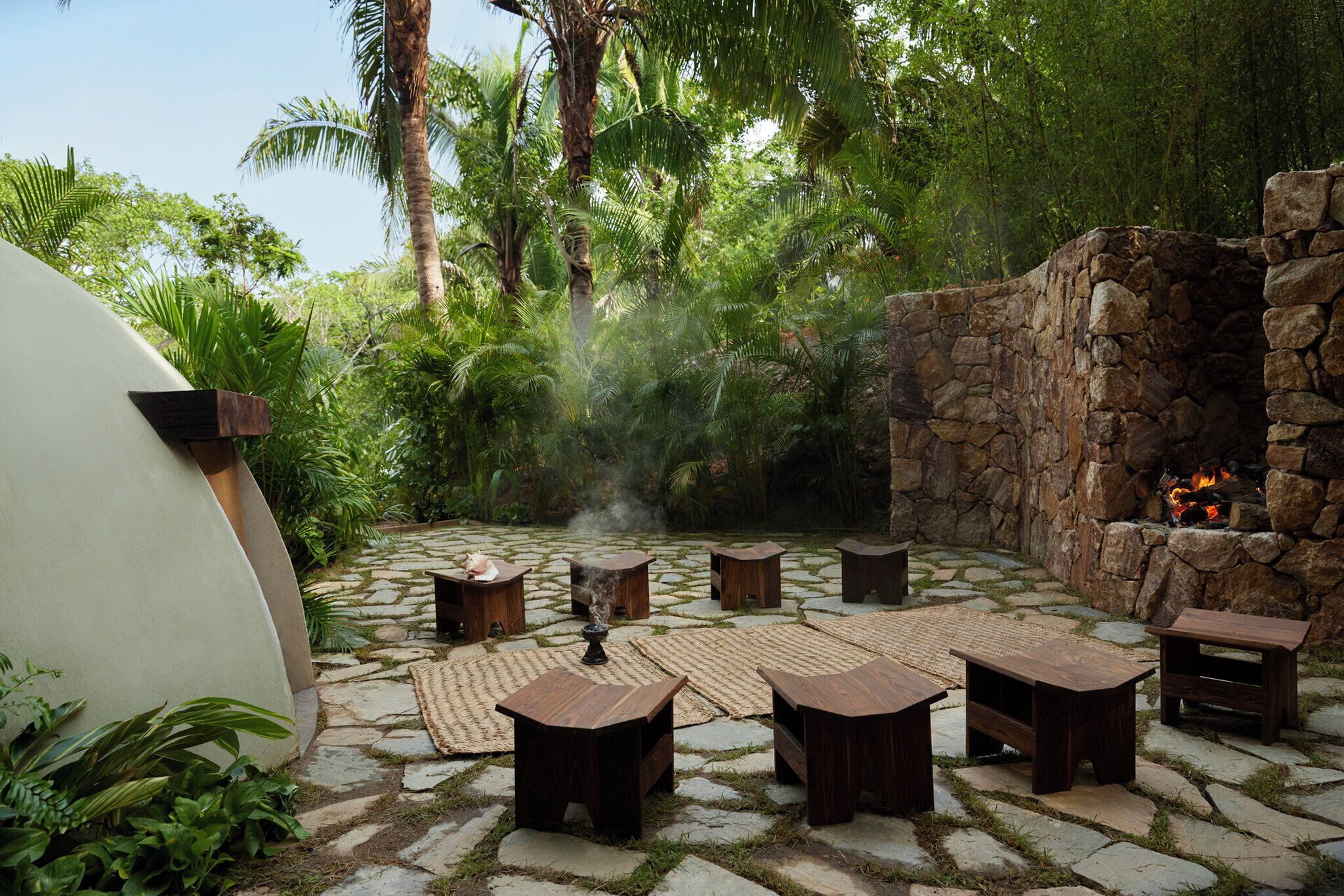 Un patio de piedra con mesas y sillas de madera, rodeado de abundante vegetación y un muro de piedra con una chimenea.