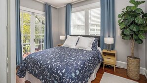 A bedroom with a bed, two bedside tables, a ceiling fan, a large window with blinds, and a potted plant.