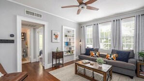 A living room with a sofa, coffee table, and a ceiling fan. There is a bookshelf and a window with curtains.